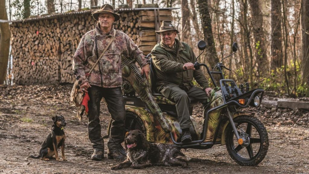 Elektrofahrzeug im Wald mit zwei Jägern und zwei Hunden